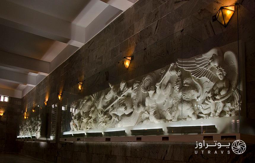 دیوار داخلی آرامگاه فردوسی که مجسمه‌های سفیدرنگ در محفظه‌های شیشه‌ای همراه با چند چراغ در بالای آن‌ها